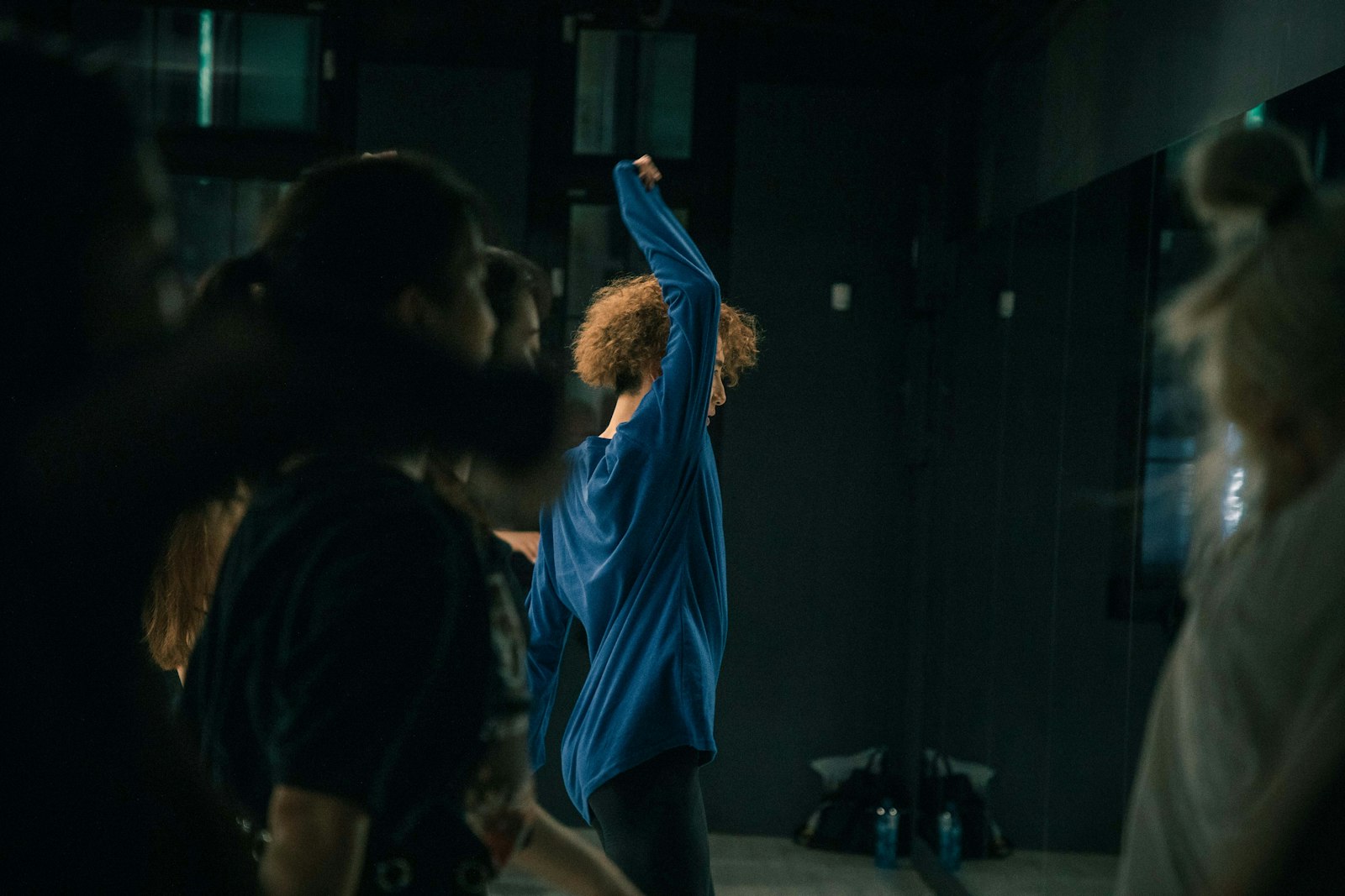 A dancer marking a move with one arm raised in a dark studio while others watch from the foreground