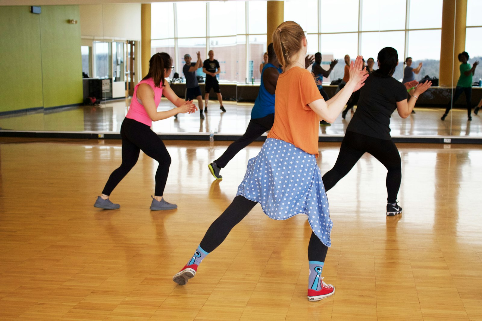An adult dance class in a mirrored studio, dancers at visibly different stages of the same move